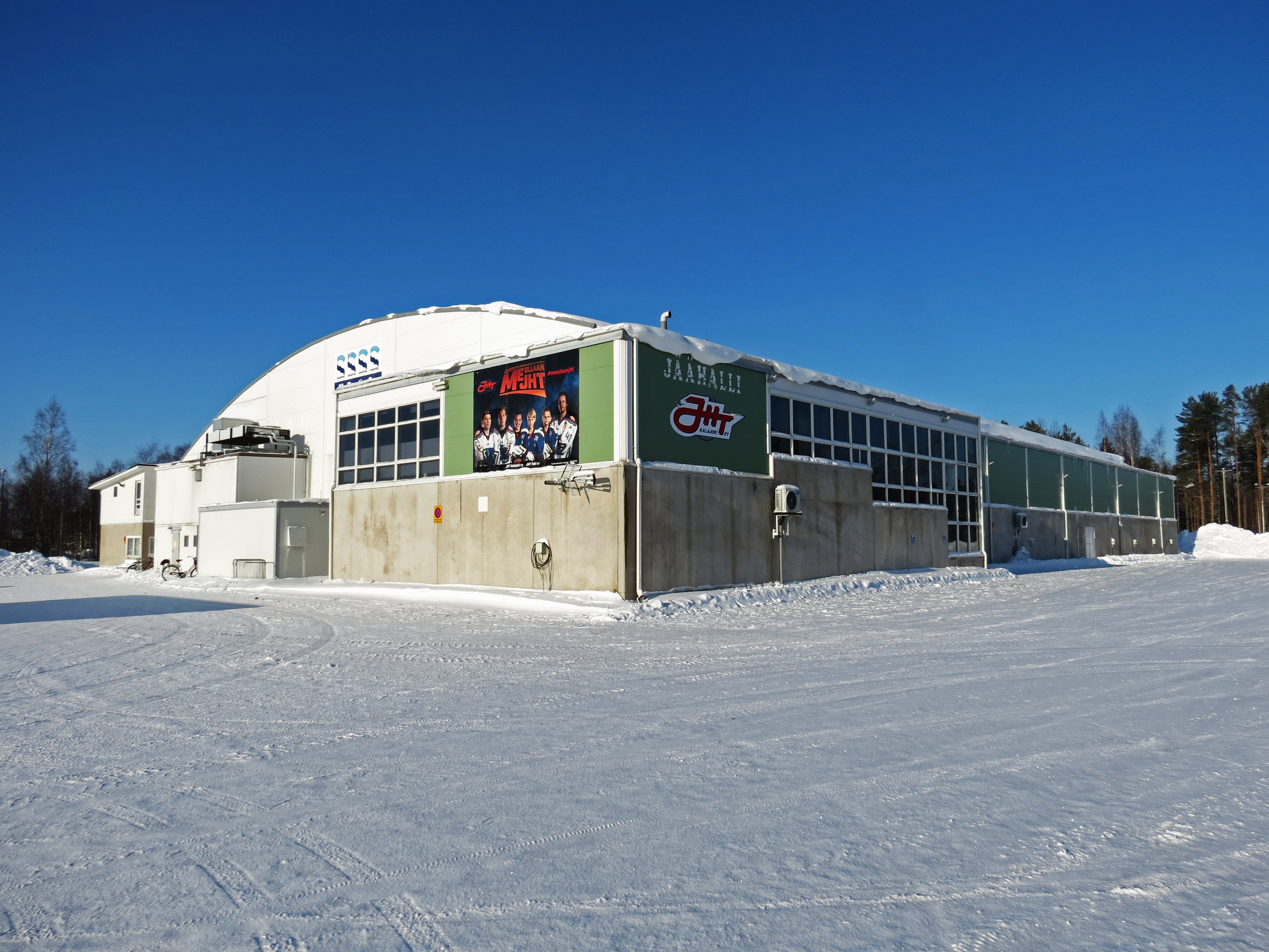Valko-vaalean vihreä iso rakennus parkkipaikan reunalla.