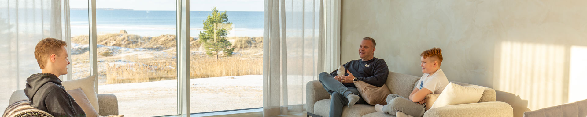 Kuvassa olohuoneessa kolme ihmistä sohvalla istumassa, merinäköala ikkunasta.