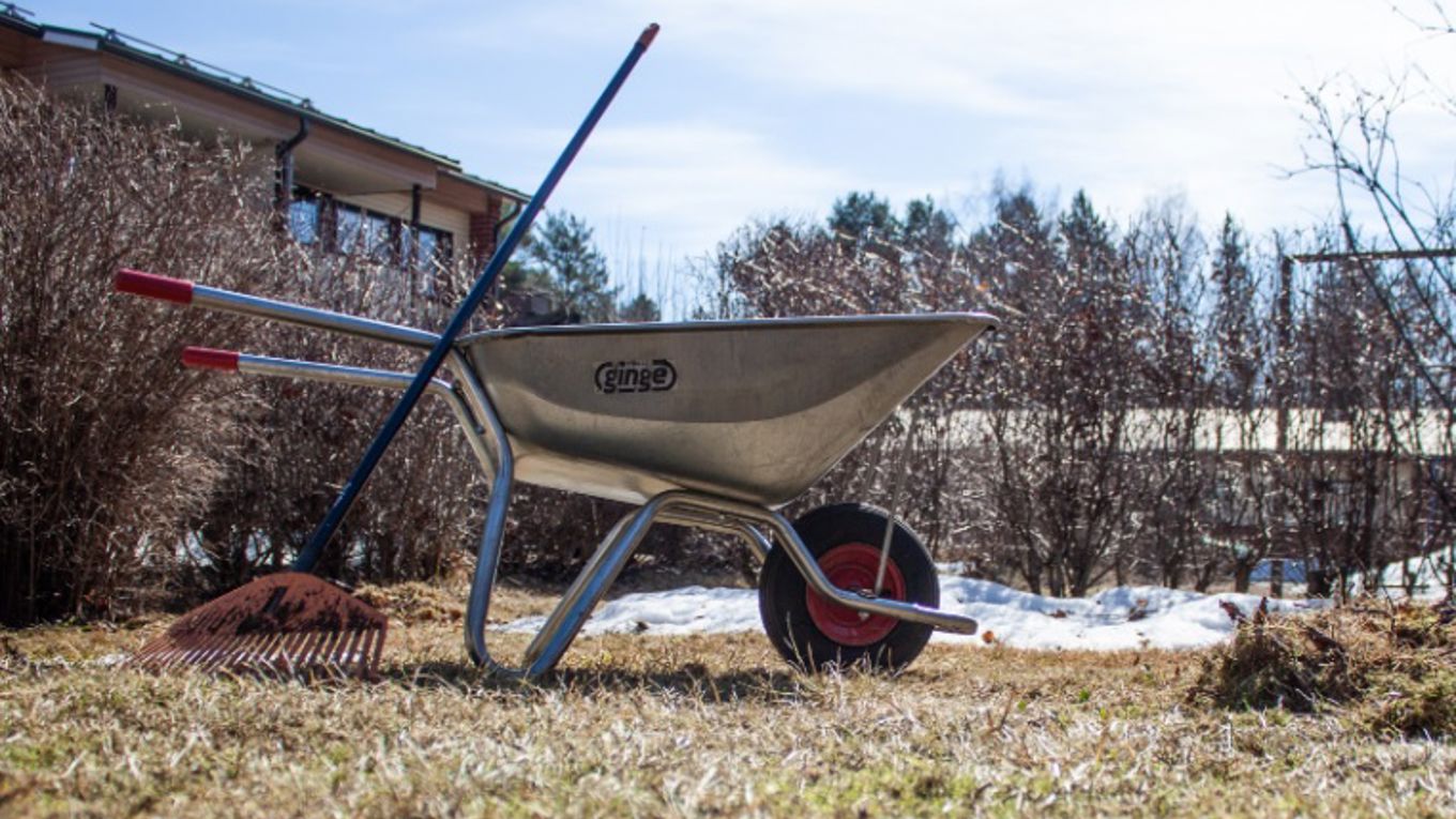 Kuvassa kevät, kottikärryt, harava, nurmikkoa, hieman lunta vielä jäljellä puskien alla