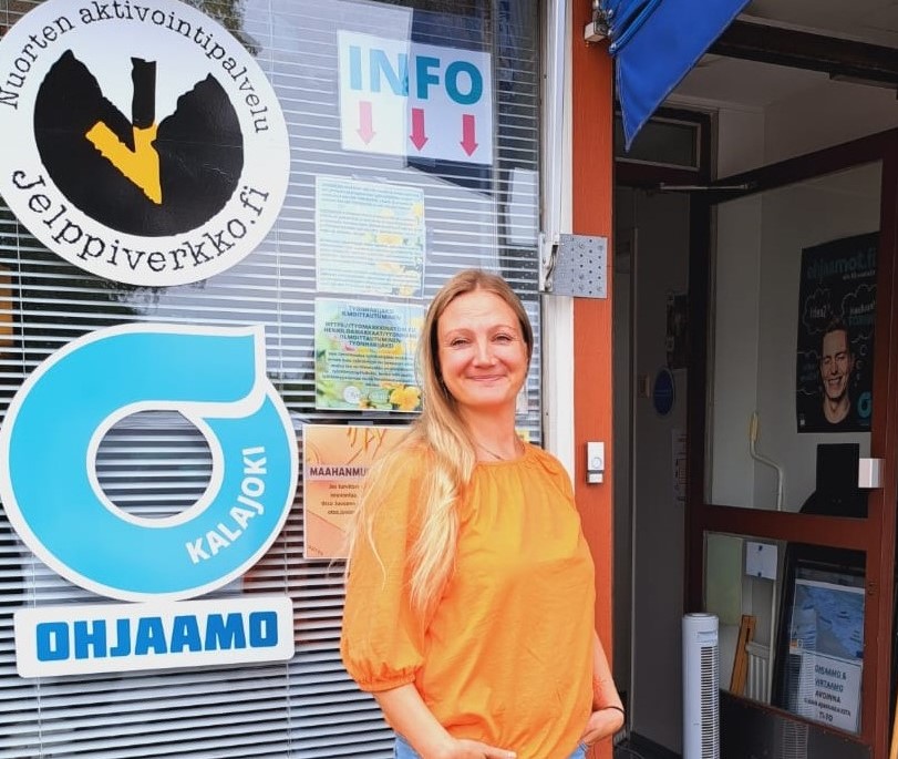 Ohjaamotyöntekijä Ohjaamon ovella. Ikkunassa Ohjaamon ja Jelppiverkon logot.