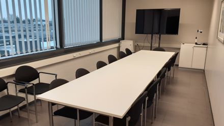 Pitkä pöytä ja 12 tuolia ympärillä. Tv. Isot ikkunat.